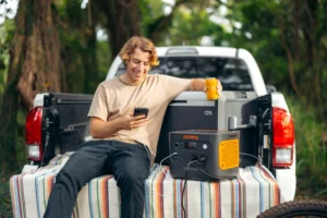 Un joven sentado en la caja de una pick-up cargando su móvil y una nevera portátil con la Jackery Explorer 1000 v2.