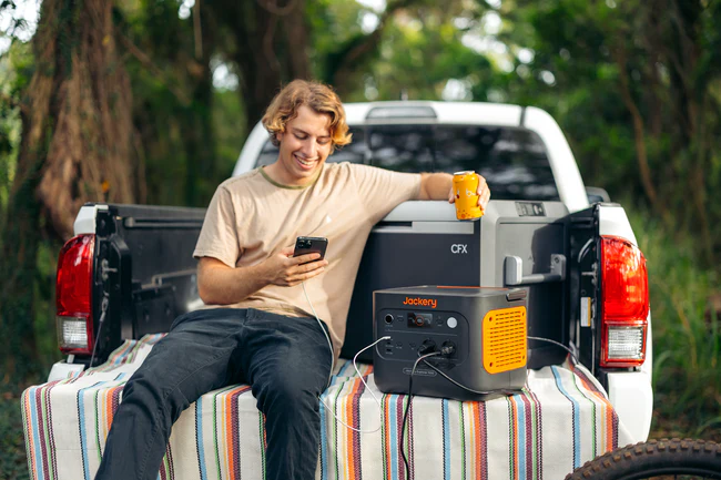 Un joven sentado en la caja de una pick-up cargando su móvil y una nevera portátil con la Jackery Explorer 1000 v2.