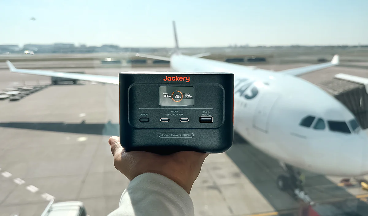 Jackery Explorer 100 Plus sostenida en la terminal de un aeropuerto con aviones de fondo.