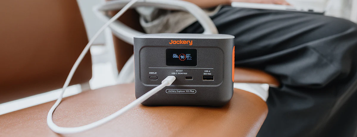 Jackery Explorer 100 Plus cargando un portátil sobre un asiento, destacando su chasis robusto.