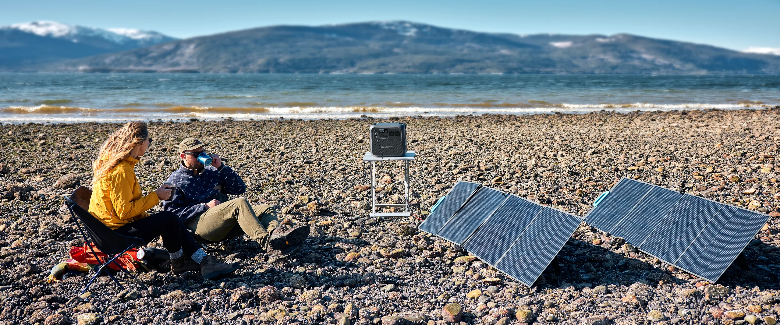 Pareja cargando su Bluetti AC180 con paneles solares plegables en una playa rocosa.