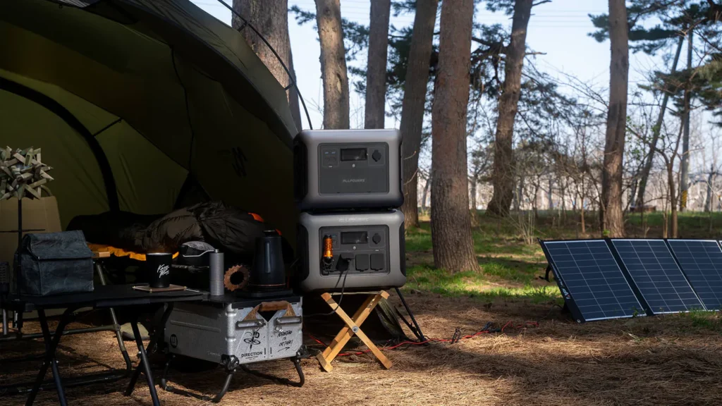 Estación de energía ALLPOWERS R1500 apilada con batería de expansión B1000 en un campamento al aire libre entre árboles, con panel solar desplegado en el suelo y mesa de camping con cafetera