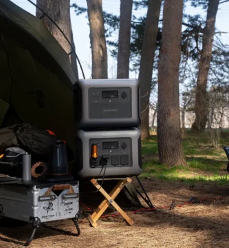 Estación de energía ALLPOWERS R1500 apilada con batería de expansión B1000 en un campamento al aire libre entre árboles, con panel solar desplegado en el suelo y mesa de camping con cafetera
