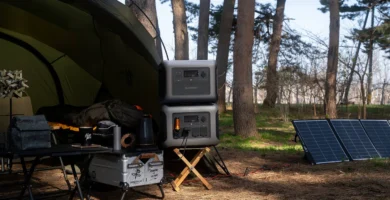 Estación de energía ALLPOWERS R1500 apilada con batería de expansión B1000 en un campamento al aire libre entre árboles, con panel solar desplegado en el suelo y mesa de camping con cafetera